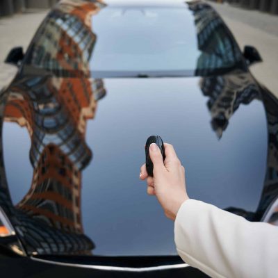 Women's hand pressing on remote control automobile alarm systems. Female arm holding car key on the background of luxure modern black auto.
